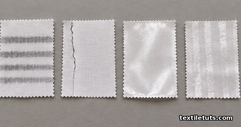 Four white cotton fabric swatches on a grey surface showing typical calendaring defects: banding, edge cracking, glazing, and uneven luster.