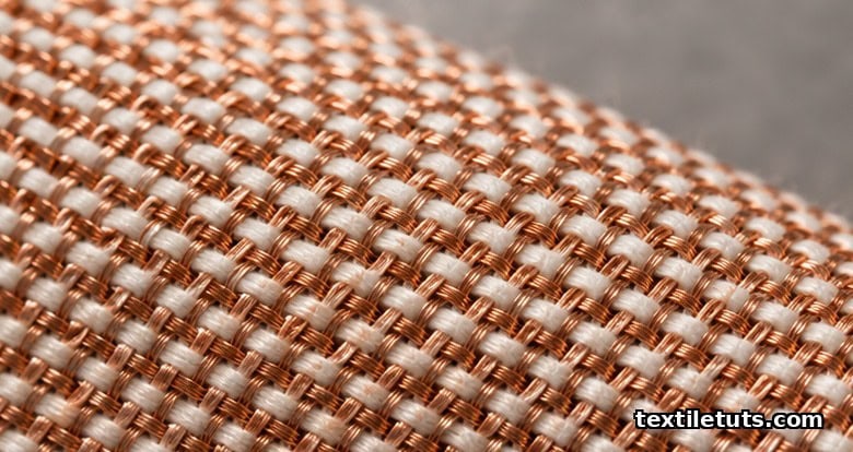 Macro close-up of a woven copper wire textile showing fine copper filaments interlaced with cotton yarns in a plain weave.