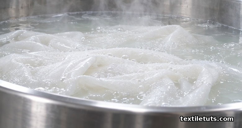 White cotton fabric submerged in a stainless steel industrial bleach bath with pale translucent liquor, small bubbles, and rising steam.