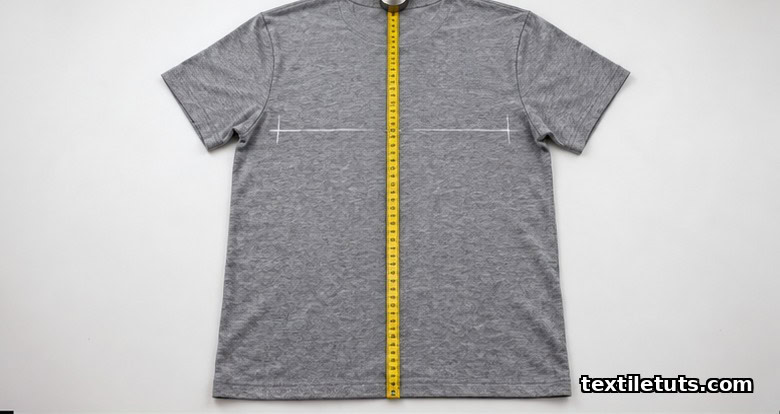 Flat crew-neck t-shirt laid on a white cutting table with a measuring tape running from high shoulder down to hem for body length.
