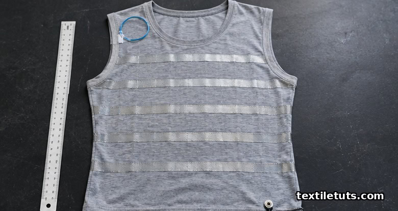 Flat-lay of a sensate monitoring undershirt showing silver-coated electrode bands, optical fiber loop, and snap connector.