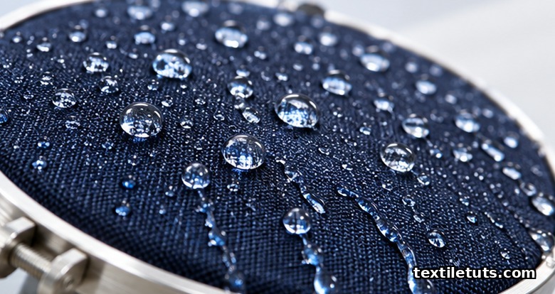 Water droplets beading on navy textile sample mounted in a metal hoop, illustrating a water-repellent finish test.